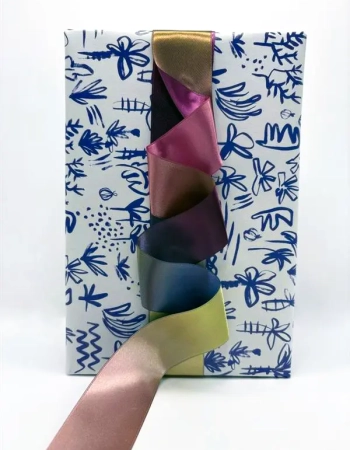Main tirant un ruban imprimé en dégradé jaune, rose, prune et bleu devant un coffret à motifs bleus — Maison Satab
