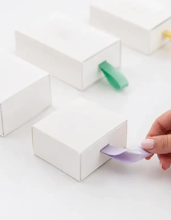 Coffrets blancs à tirettes en ruban satin mauve, vert et jaune, une main tirant la languette — Maison Satab